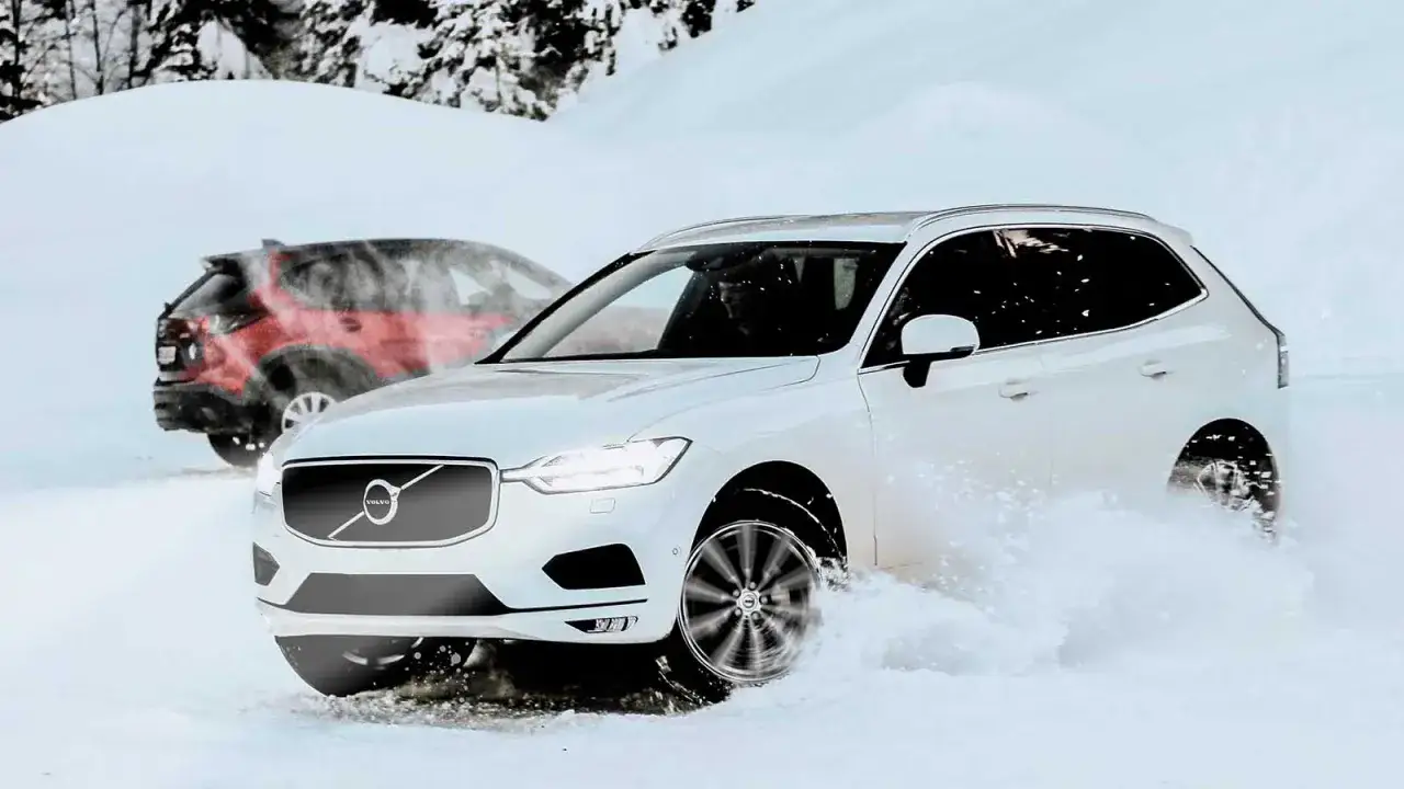 Vit Volvo k&ouml;r genom sn&ouml;, en r&ouml;d bil syns i bakgrunden. AWD betyder trygghet i vinterv&auml;glag.