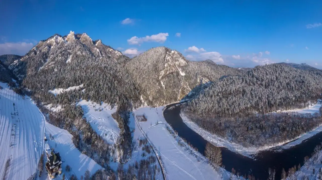 Zimowe Pieniny, rzeka Dunajec meandruje wśród ośnieżonych lasów. W oddali skaliste szczyty. W dole widać samotny dworek.