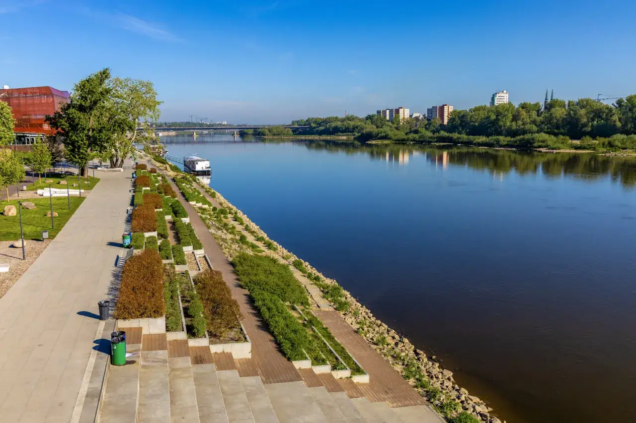 Nowoczesne miasto nad Wisłą z bulwarami, zielenią i budynkami w tle.