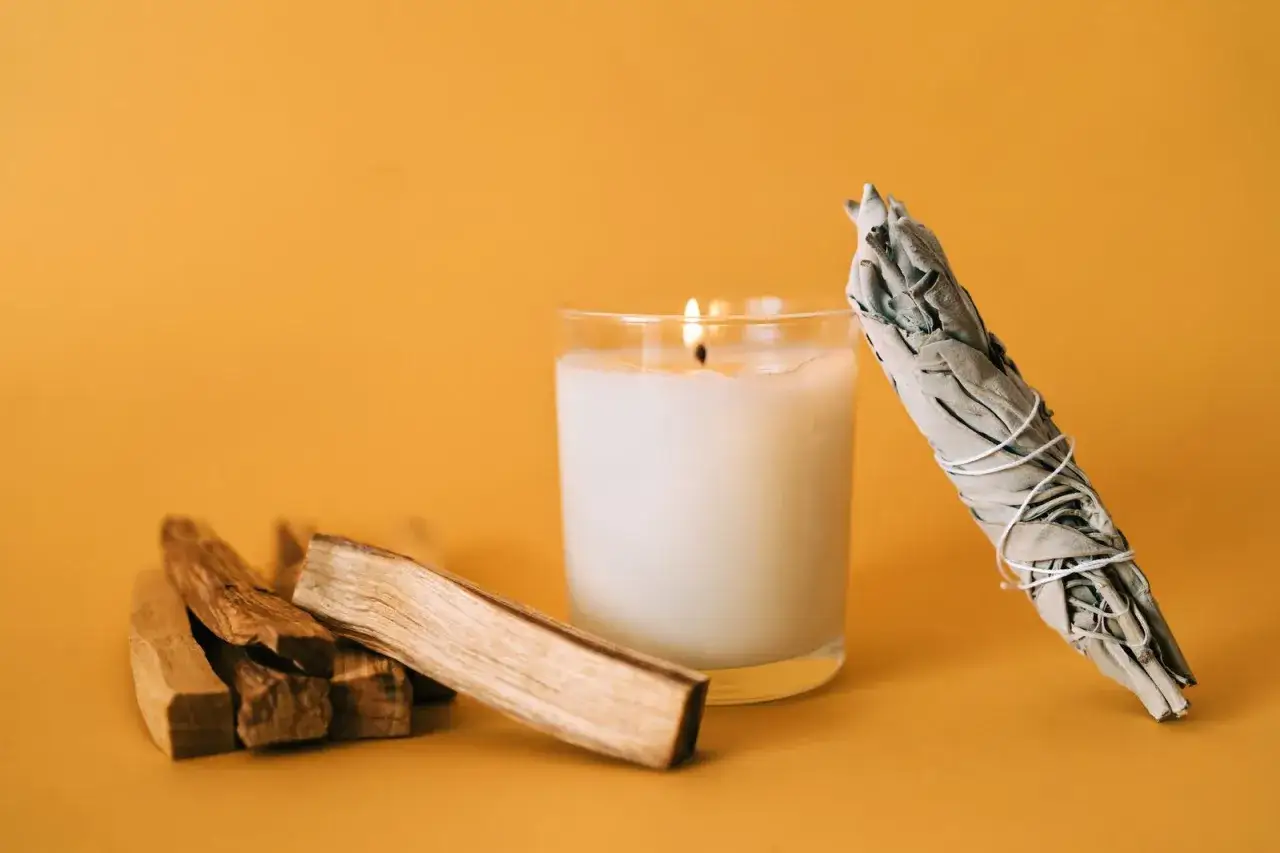 Palo santo para que sirve: madera arom&aacute;tica, vela encendida y manojo de salvia blanca sobre fondo naranja.