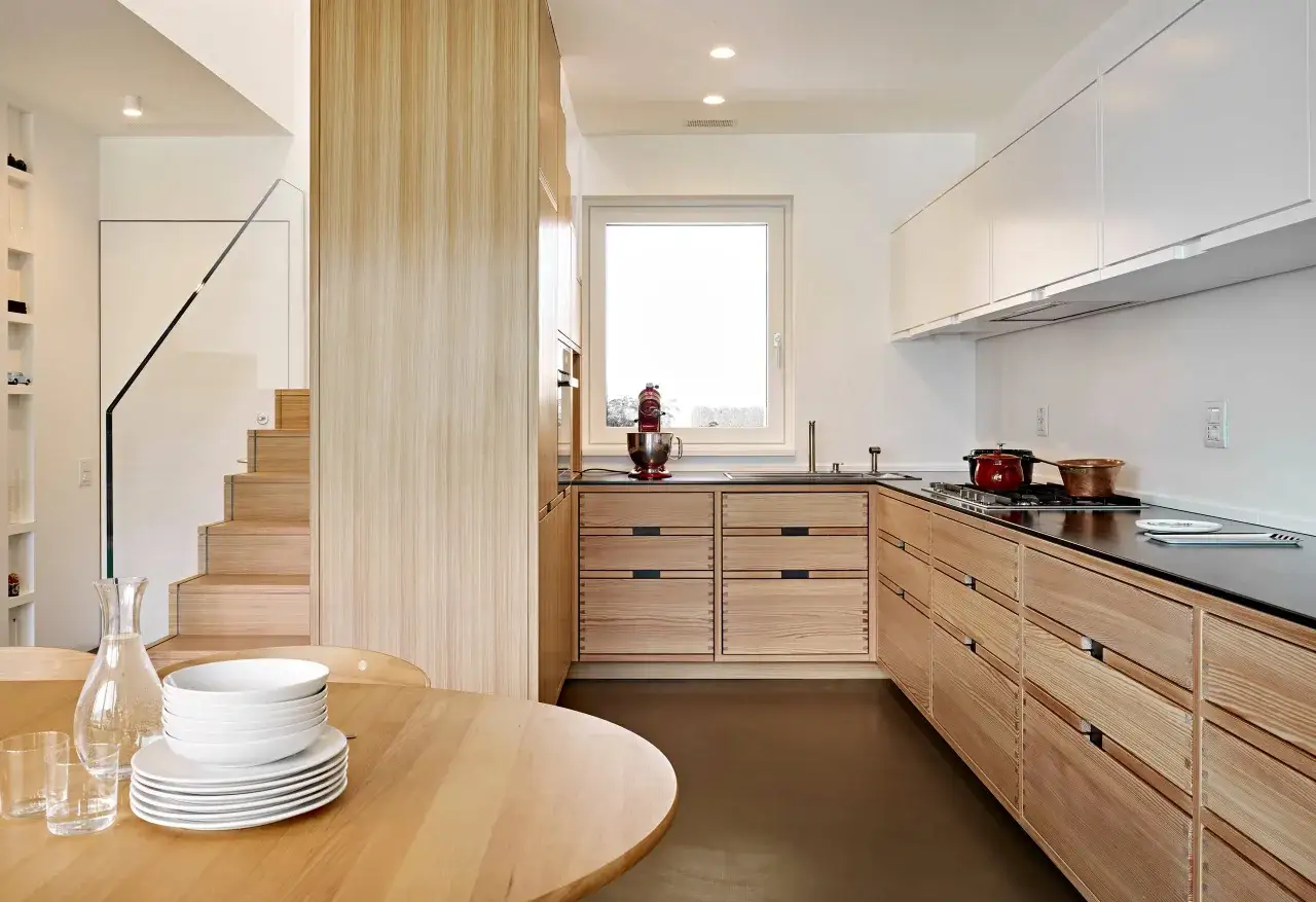 Cucina zen con mobili in legno chiaro, piano di lavoro scuro e scala in vetro. Atmosfera minimalista e ordinata.