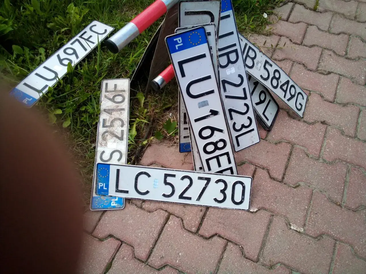 Zgubiona tablica rejestracyjna LC 52730 leży wśród innych tablic, jak LU 697FC i S 2516F, na chodniku.