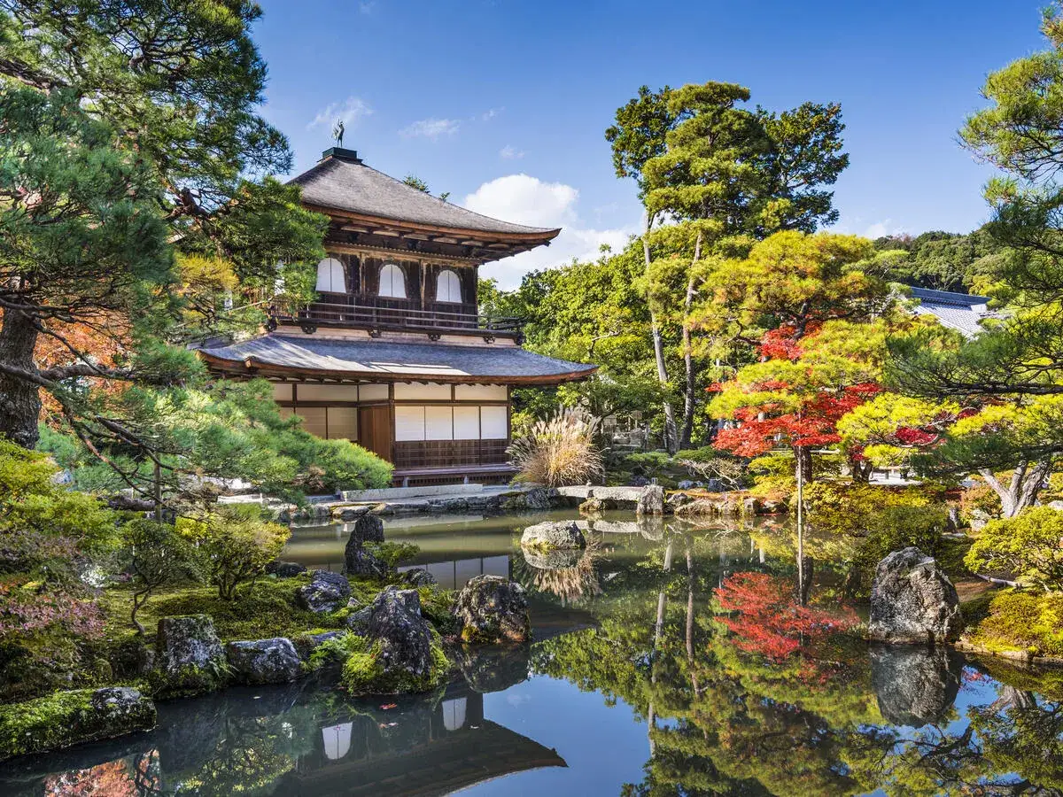 Ein friedlicher asiatischer Garten mit einem traditionellen Gebäude, einem Teich und bunten Herbstbäumen, die sich im Wasser spiegeln.