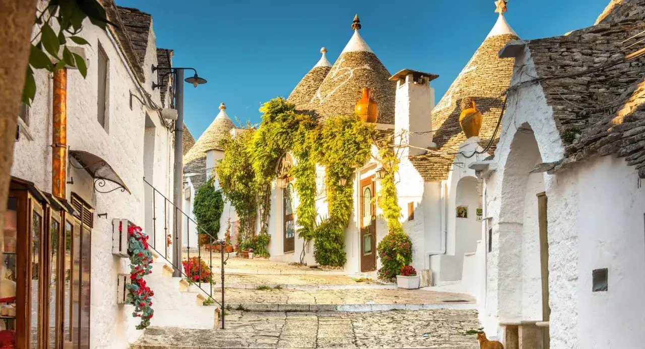 Un vicolo fiorito tra i trulli, nel tacco dell'Italia. Case bianche con tetti conici in pietra, un gatto arancione passeggia.