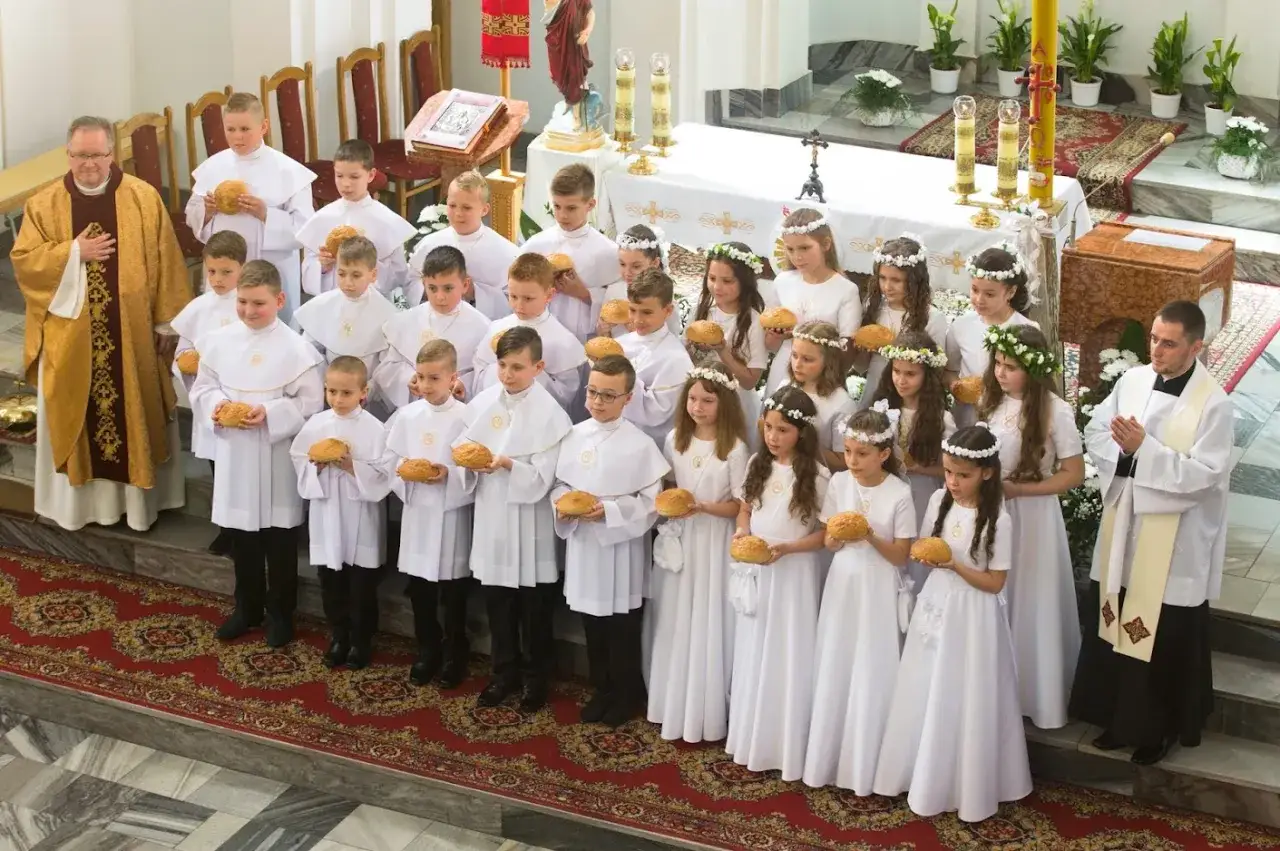 Dzieci w białych strojach, z wiankami na głowach, trzymają chlebki podczas uroczystości komunii świętej. Obok kapłan w złotej i białej szacie.