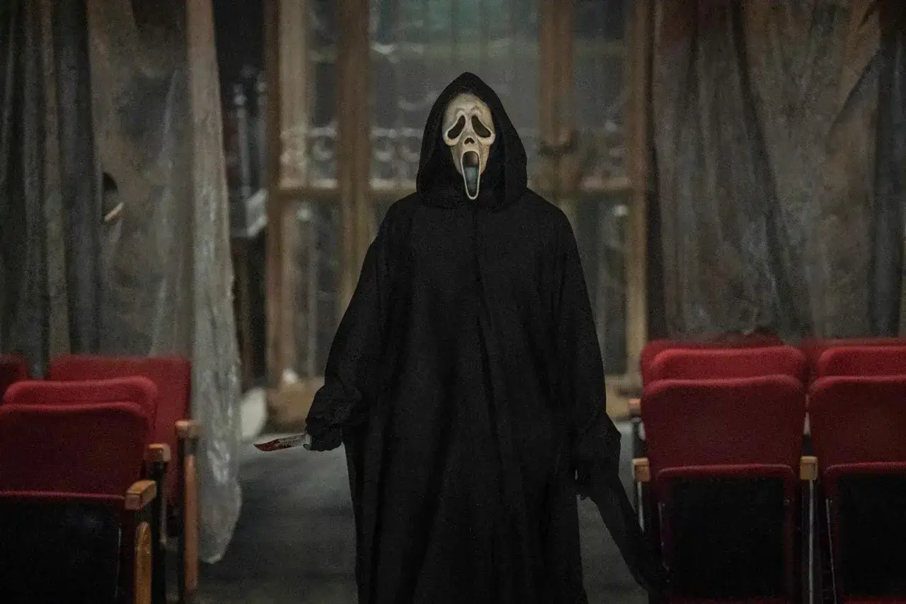 Postać w masce Ghostface z nożem, w czarnej szacie, stoi w pustej sali kinowej. Klasyczny slasher horror.