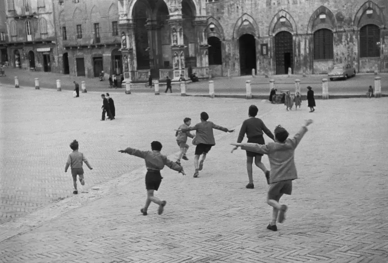 Bambini corrono in una piazza, un'istantanea che ricorda le cartier bresson foto famose. L'architettura storica fa da sfondo alla loro gioia.