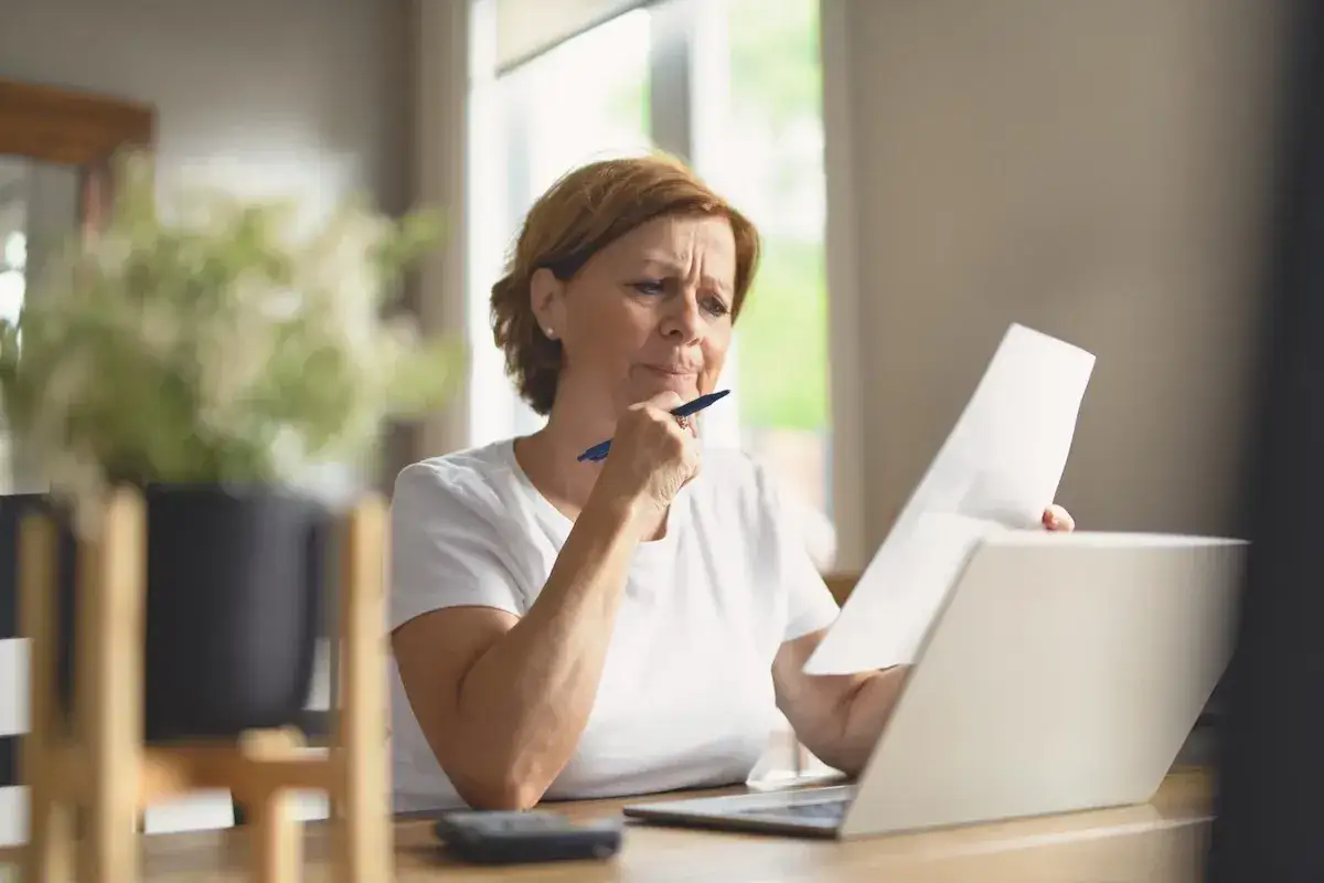 Donna pensierosa con fogli e laptop, forse gestisce le finanze del suo immobile in comproprietà abitato da un solo proprietario.