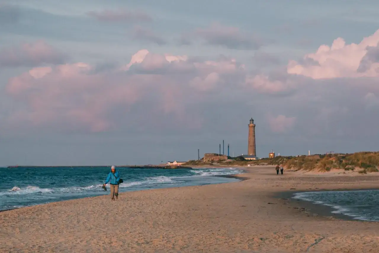 En person promenerar på en sandstrand med vågor som slår in. I bakgrunden syns skagens fyr och en molnig himmel i rosa nyanser.