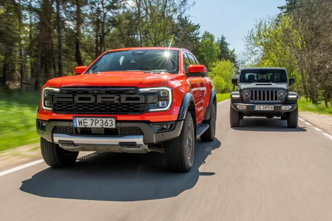 Pomarańczowy Ford Ranger Raptor i szary Jeep jadą drogą wśród drzew.