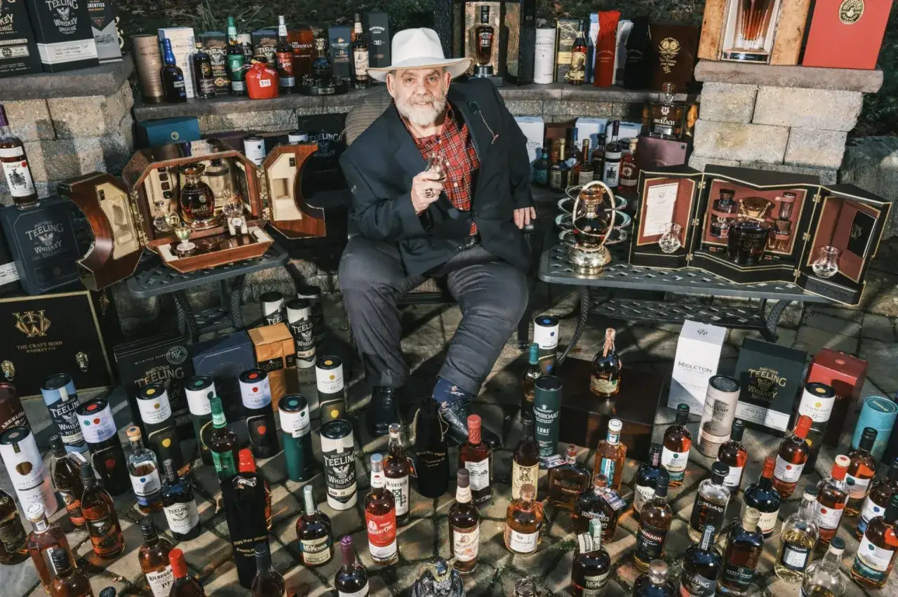 Mężczyzna w kapeluszu siedzi wśr&oacute;d kolekcji drogich whisky, trzymając kieliszek.