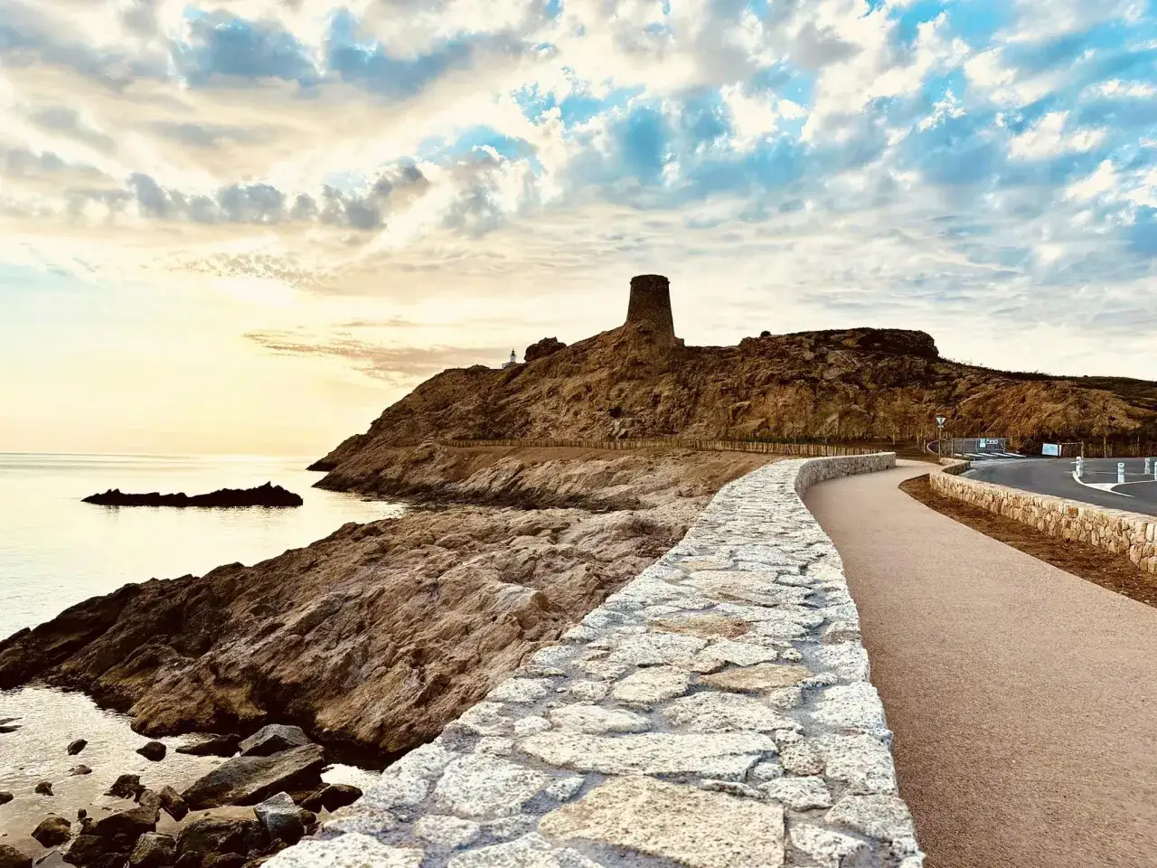 Chemin longeant une **porte de pierre** menant à une tour sur une colline rocheuse, avec la mer et un ciel nuageux au coucher du soleil.