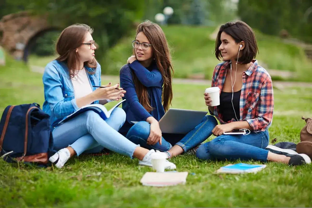 Trzy studentki dyskutują o tym, jakie studia po humanie wybrać, siedząc na trawie z książkami i laptopem.