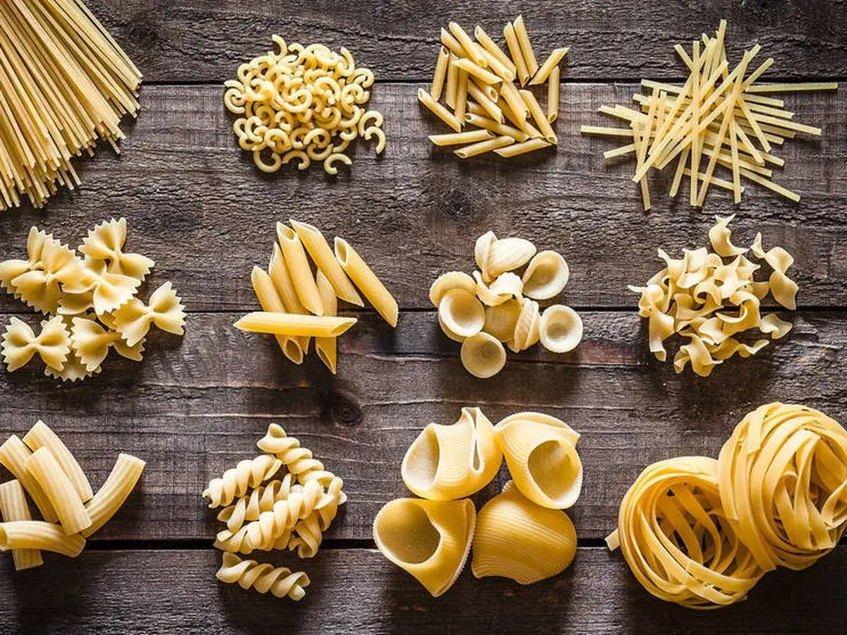 Variedad de pasta sin huevo: espagueti, farfalle, penne, orecchiette, fusilli, rigatoni, fettuccine y más, dispuestas sobre madera.