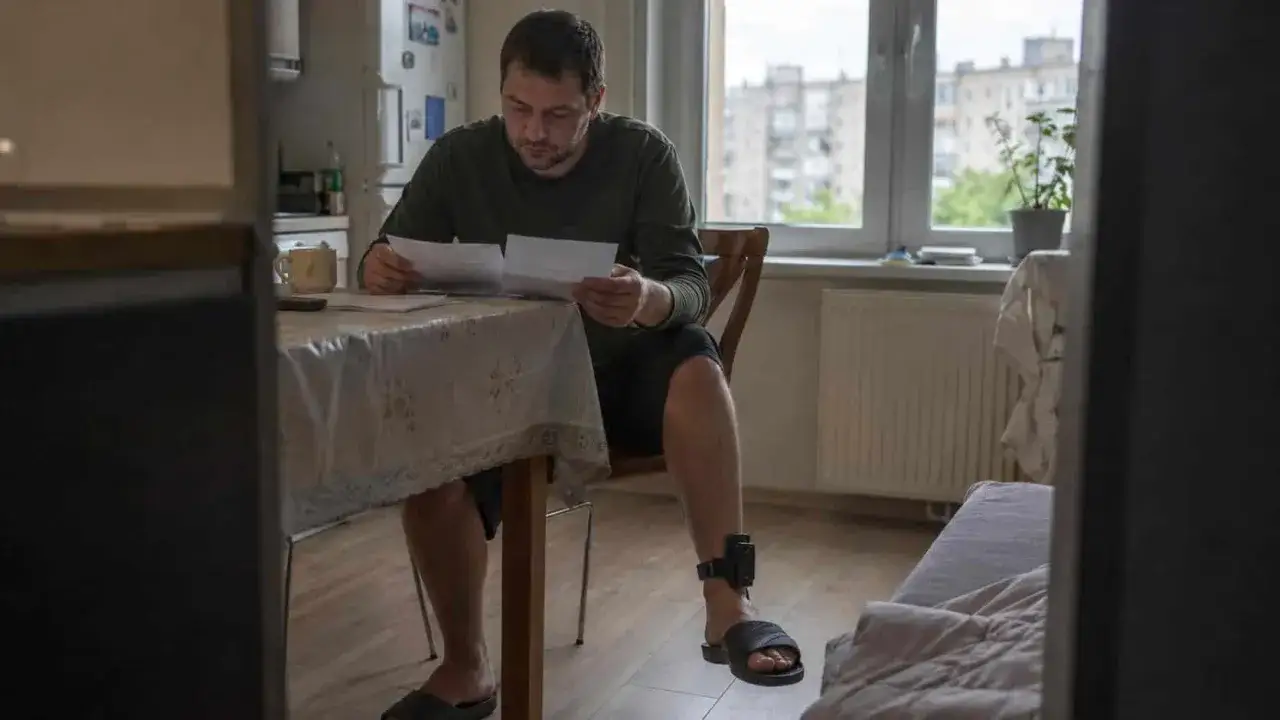 Mężczyzna z opaską na nodze, symbolizującą zatrudnienie pracownika z dozorem elektronicznym, analizuje dokumenty przy stole.