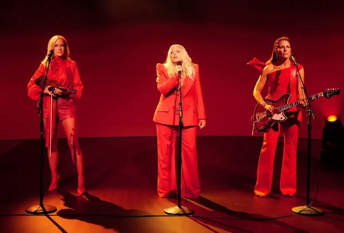 Three women in red outfits perform on stage. The woman on the left holds a violin, the center woman sings into a microphone, and the woman on the right plays a guitar. This is the Chicks setlist.