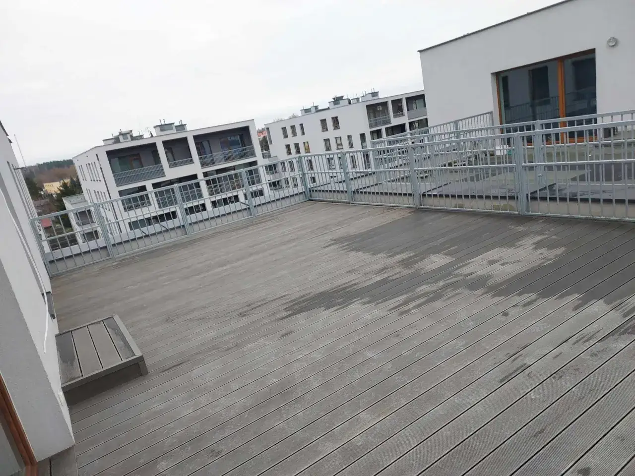 Szeroki, drewniany taras na dachu z metalową balustradą, otoczony nowoczesnymi budynkami mieszkalnymi.