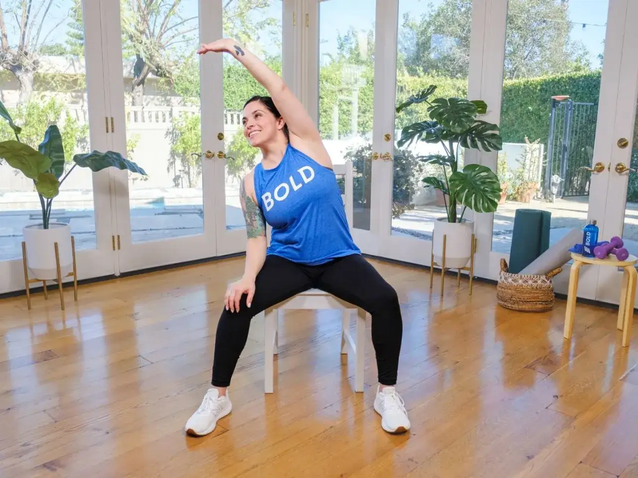 Femme souriante faisant des exercices de pilates gratuit pour seniors, assise sur une chaise, bras lev&eacute;.