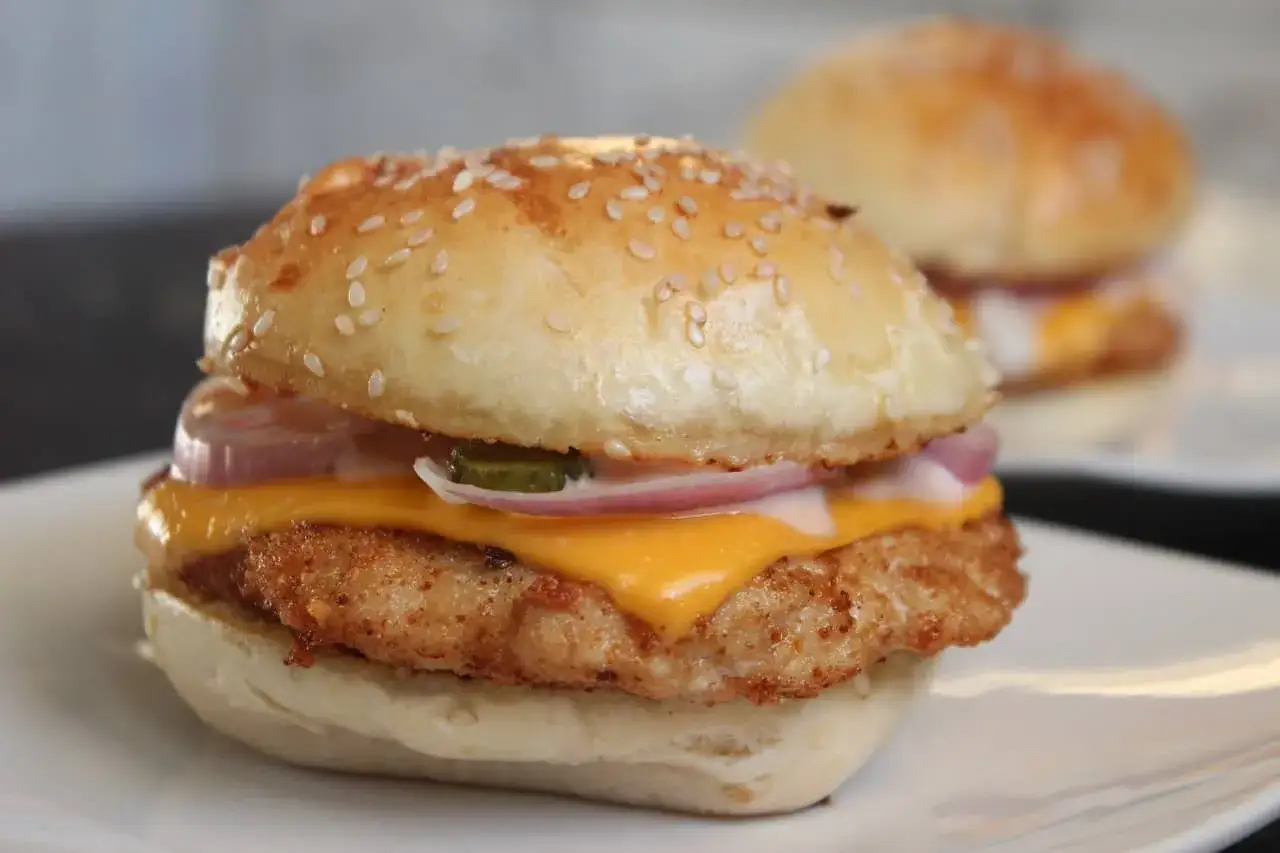 Deliciosa hamburguesa de pollo con queso derretido, cebolla morada y pepinillos, servida en un pan con semillas de s&eacute;samo.
