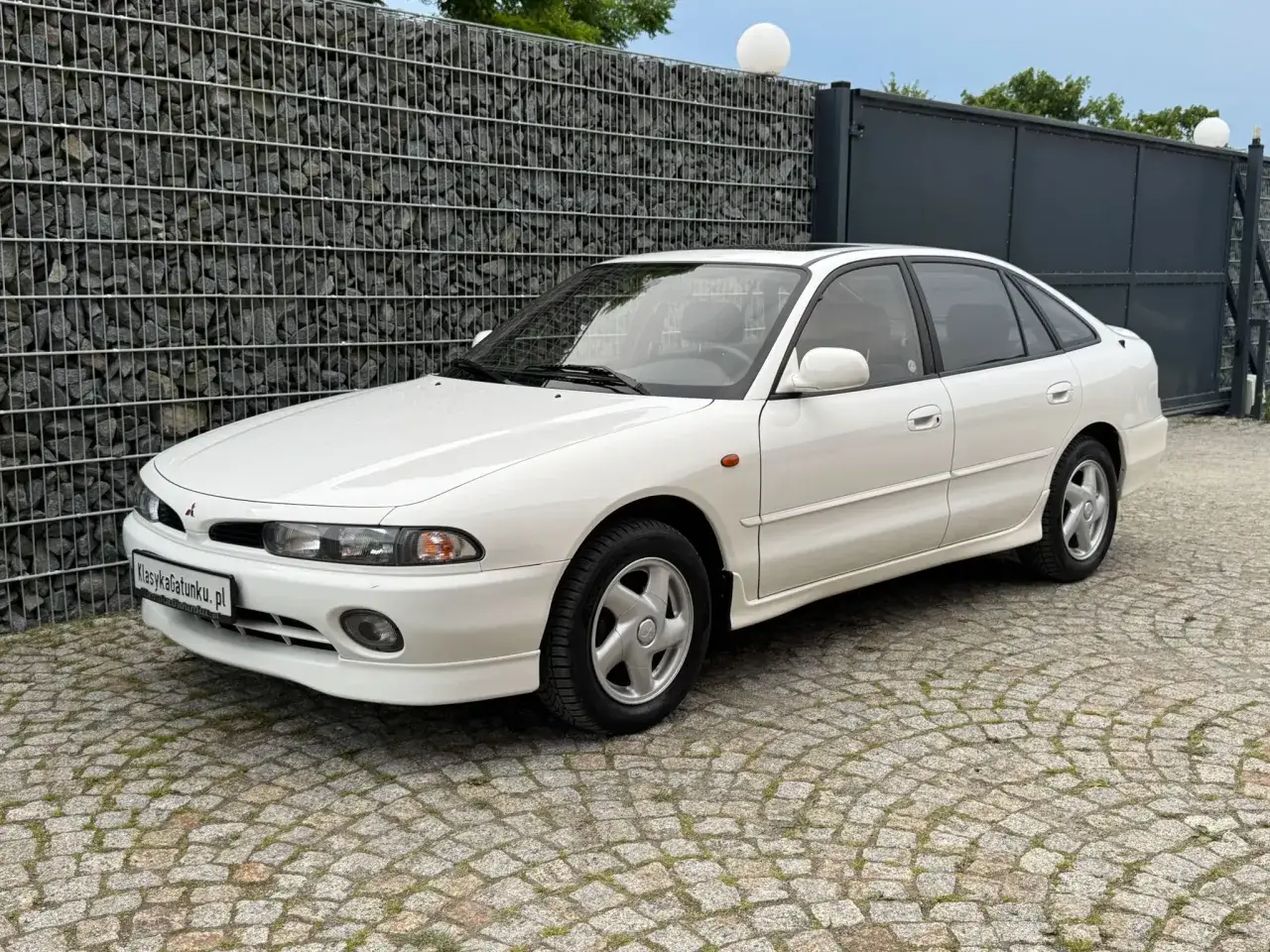 Biały, galant sedan z lat 90. stoi na brukowanym podjeździe przed kamiennym murem i nowoczesną bramą.
