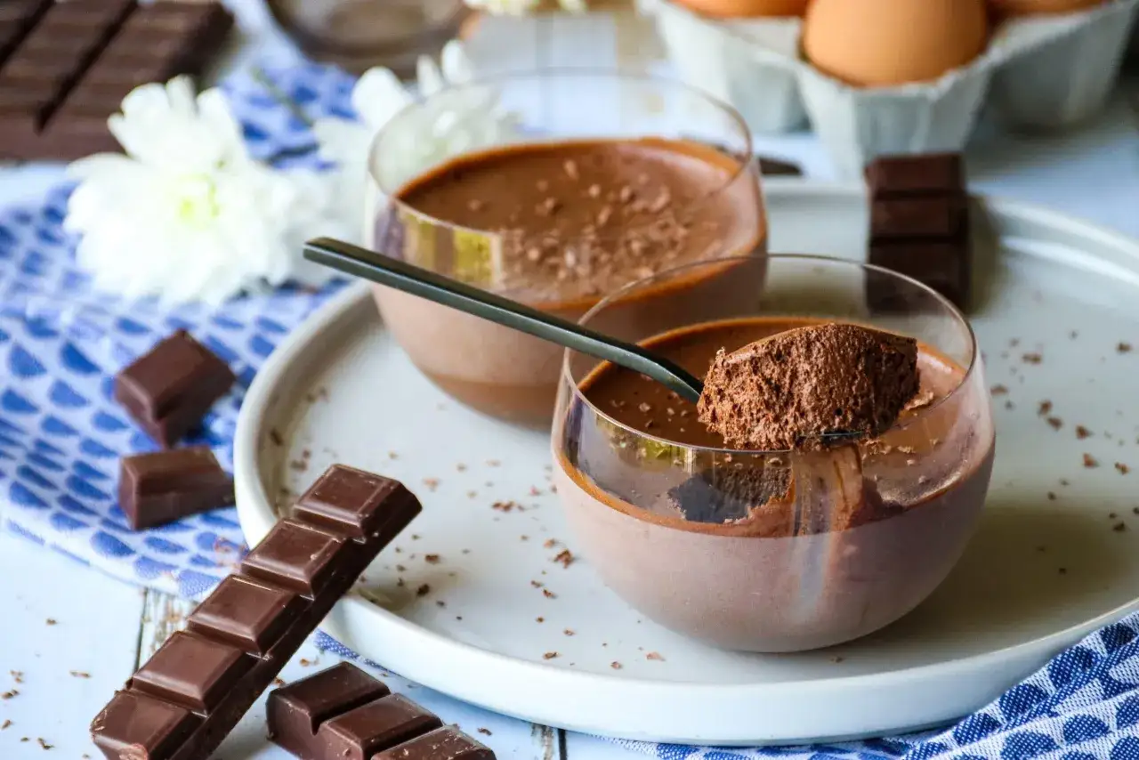 Deux coupes de mousse au chocolat skyr, garnies de cacao, avec des carrés de chocolat et des fleurs blanches en arrière-plan.