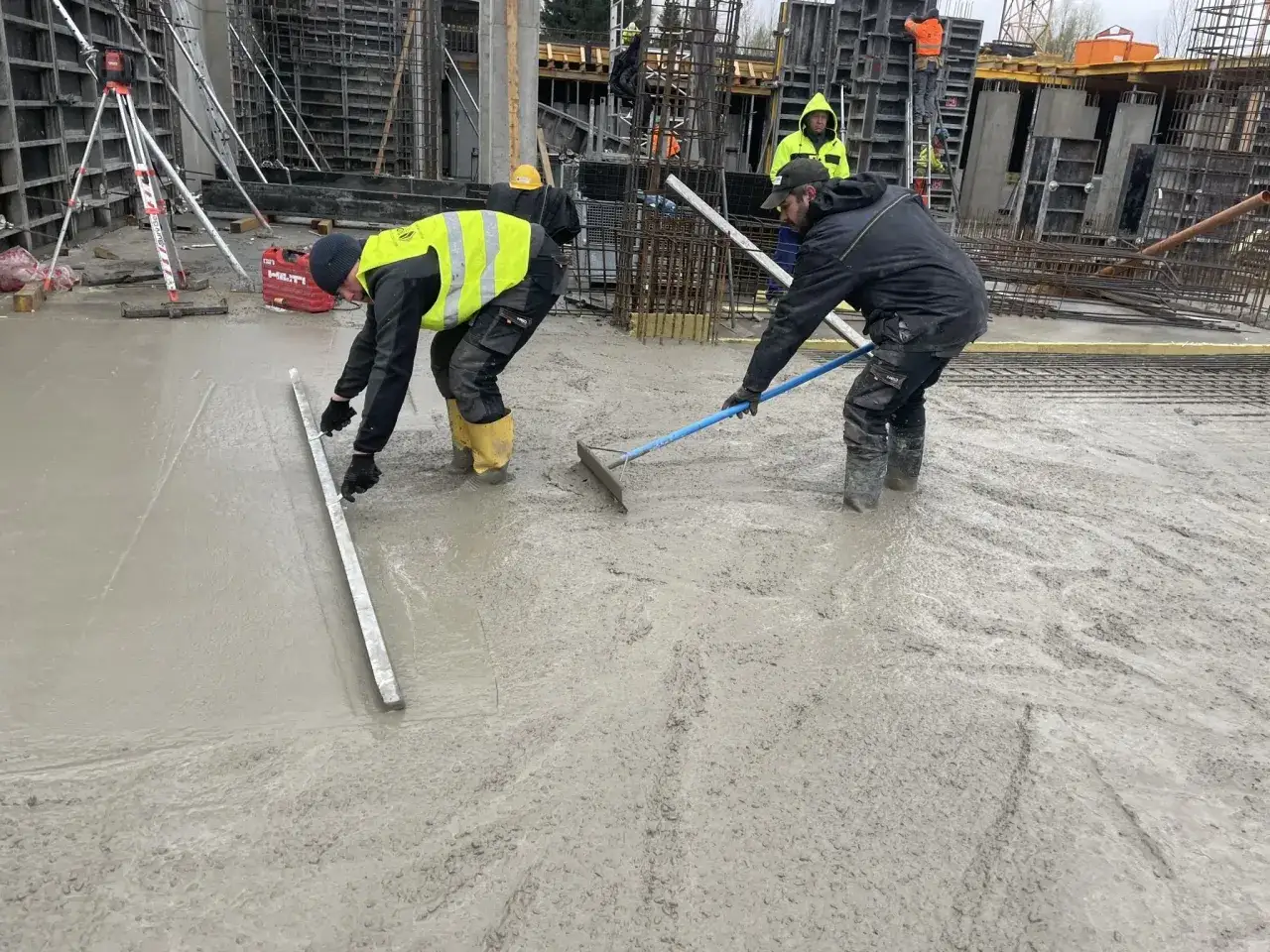 Pracownicy na budowie wygładzają świeży beton. Jeden z nich używa łaty, drugi grabi. To ważny etap prac, zanim beton stwardnieje, jak styrobeton.