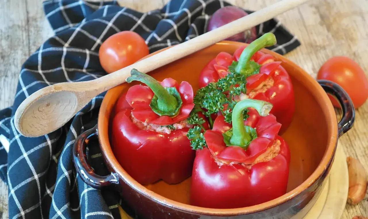 Trois poivrons farcis rouges, garnis de persil, dans un plat en terre cuite. Une cuillère en bois et des tomates sont à proximité.