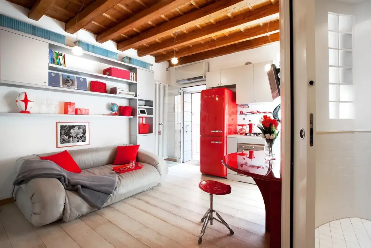 Arredare monolocale con stile: divano grigio, cuscini rossi, frigo Smeg rosso acceso e travi a vista.