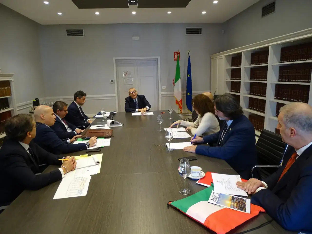 Riunione di lavoro in Italia, con bandiere italiana ed europea. Persone sedute a un tavolo, intenti a discutere e firmare documenti.