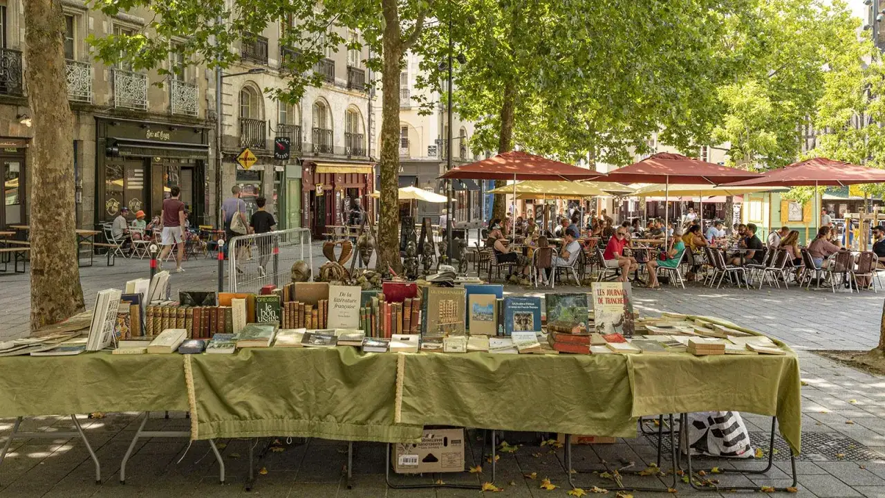 Ein Buchstand mit alten B&uuml;chern auf einem sonnigen Platz in Rennes, Frankreich. Im Hintergrund sitzen Menschen in einem Stra&szlig;encaf&eacute;.