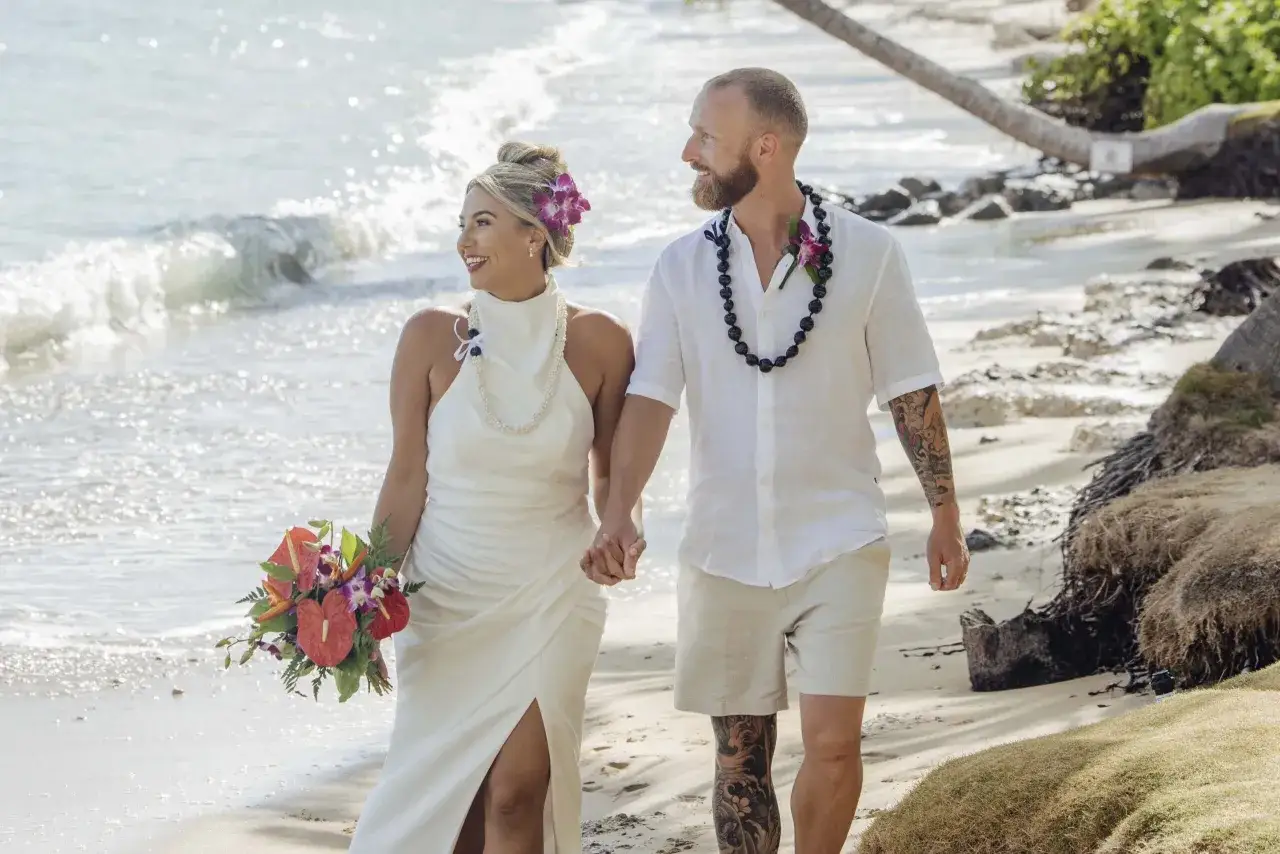 Newlyweds stroll on a beach, showcasing perfect what to wear to beach wedding attire: a flowing white gown and a linen shirt with shorts.