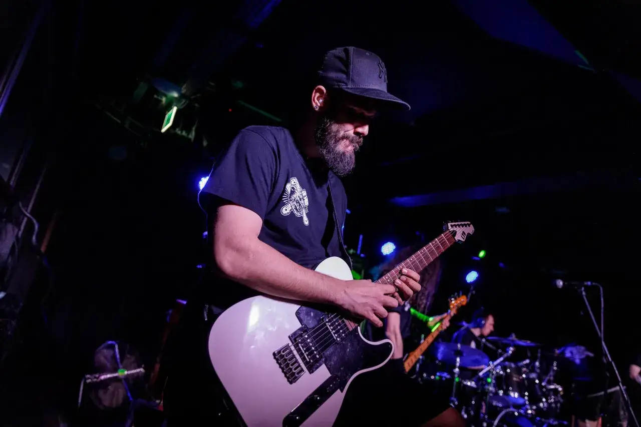 Mężczyzna z brodą i czapką z daszkiem gra na białej gitarze elektrycznej. Brzmienie jak siberian meat grinder.