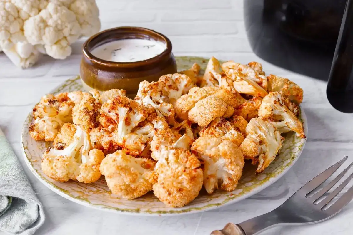 Coliflor airfryer crujiente y dorada, servida en un plato con un tazón de salsa cremosa.