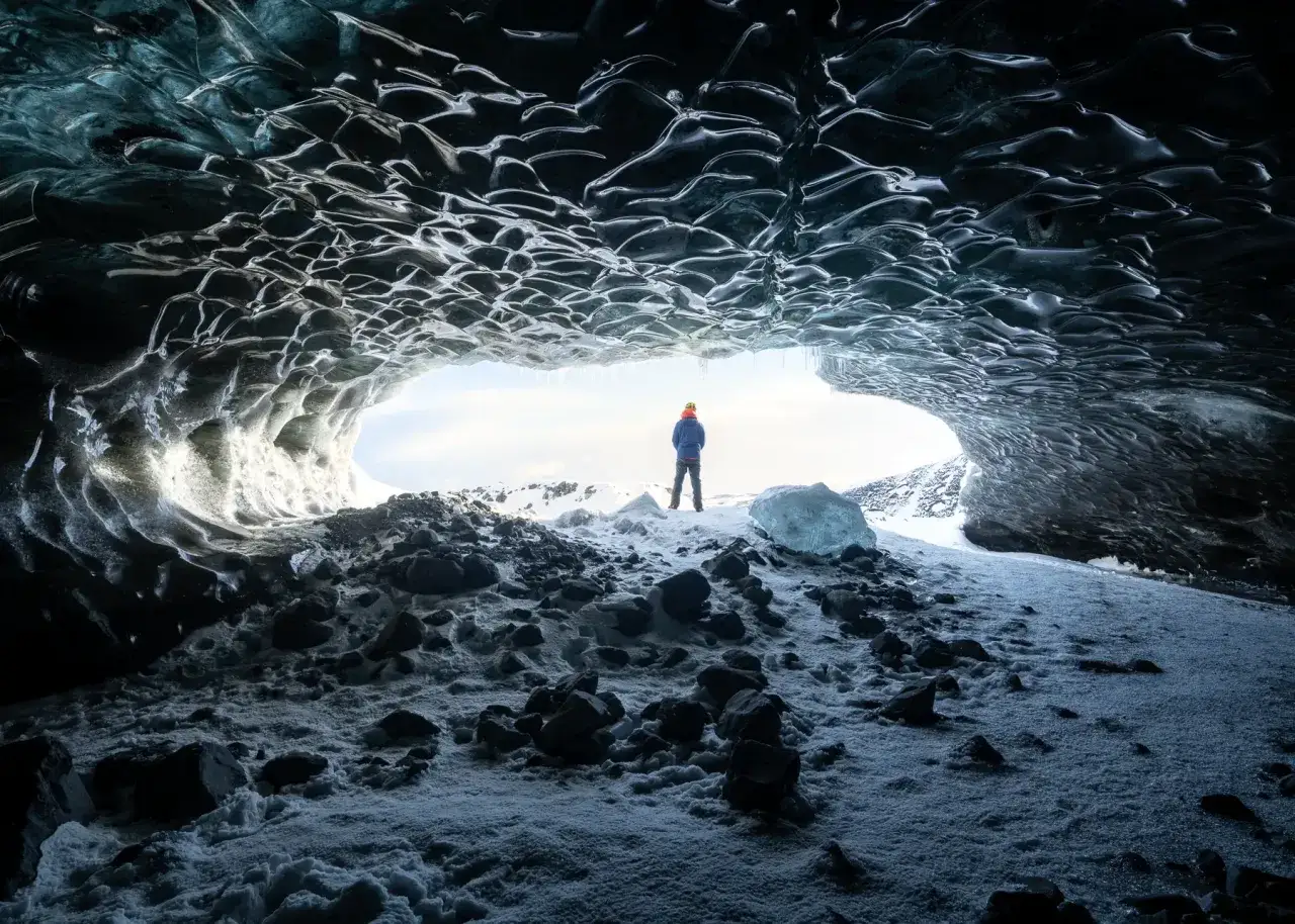 Persona con gorro naranja y chaqueta azul se asoma desde una cueva de hielo, rodeada de rocas y nieve. Bonitas fotos en la nieve de un paisaje helado.