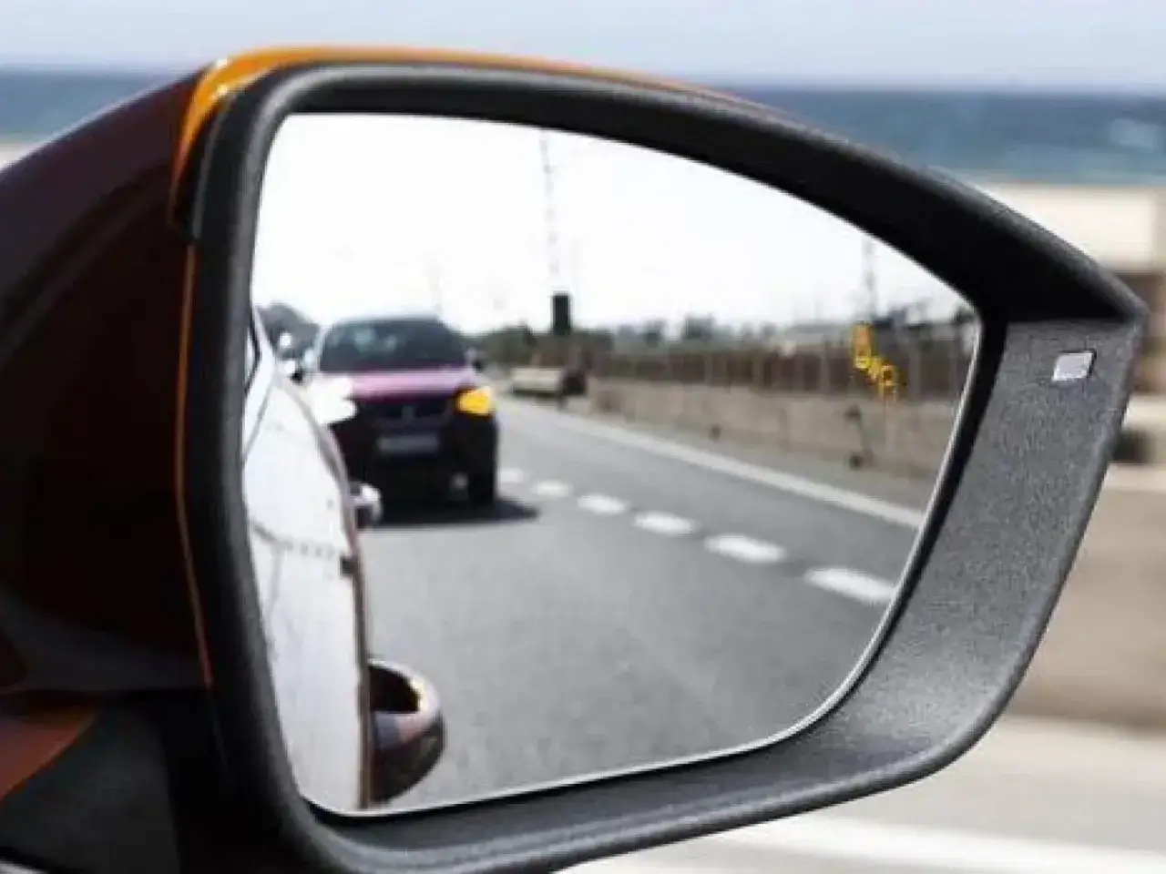 El retrovisor lateral es obligatorio en coche. Refleja un coche morado en la carretera, con un punto amarillo de alerta de punto ciego.