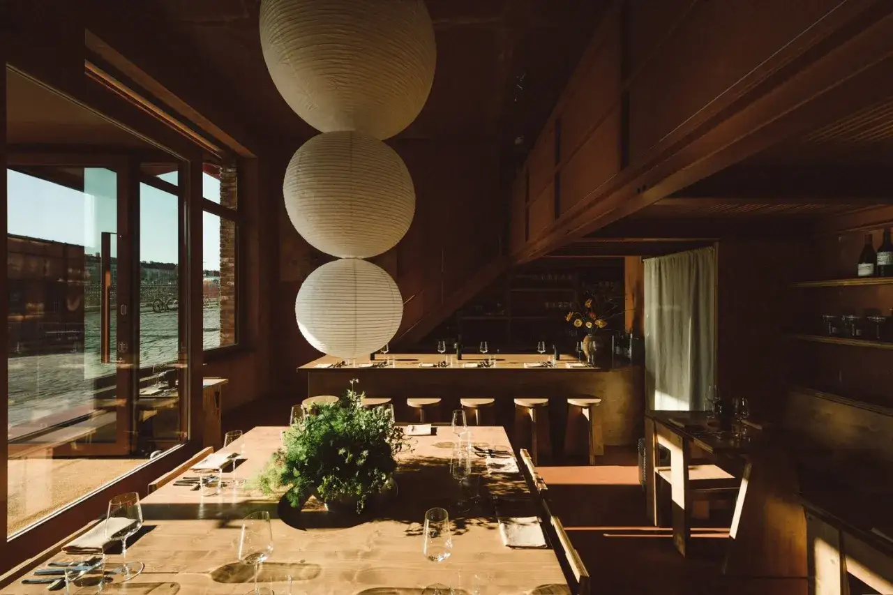 Gem&uuml;tliches Interieur eines Restaurants in **Aerde Berlin** mit Holztischen, gro&szlig;en Papierlampen und Blick auf das Wasser.