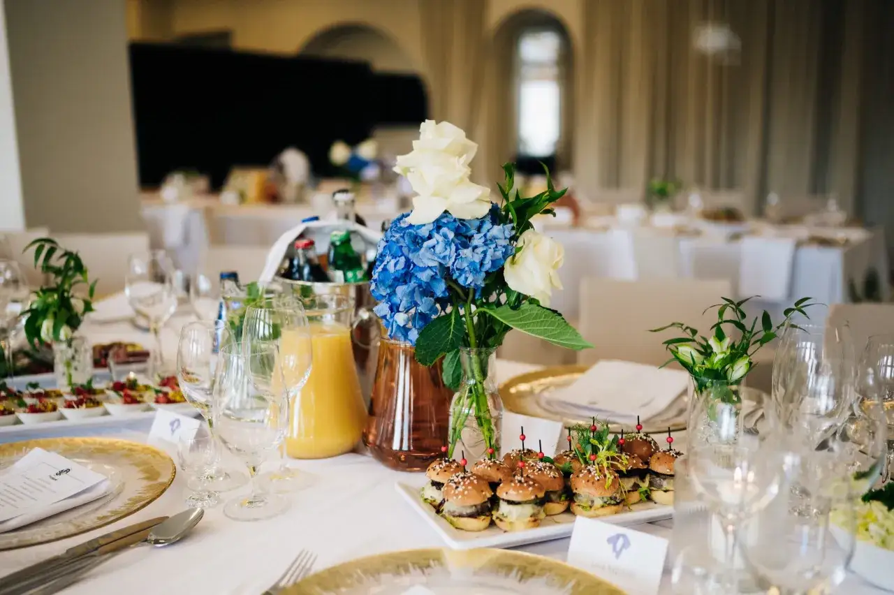Uroczyste chrzciny w restauracji: st&oacute;ł z mini burgerami, sokiem, kwiatami i elegancką zastawą.