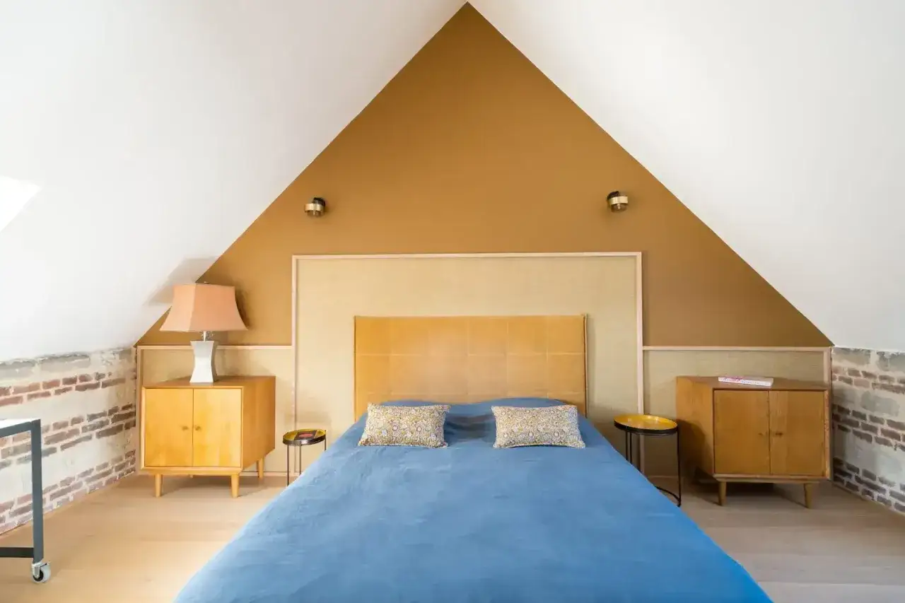 Chambre sous pente avec mur en **couleur ocre** et lit bleu. Tables de chevet en bois, lampes et murs en briques apparentes.