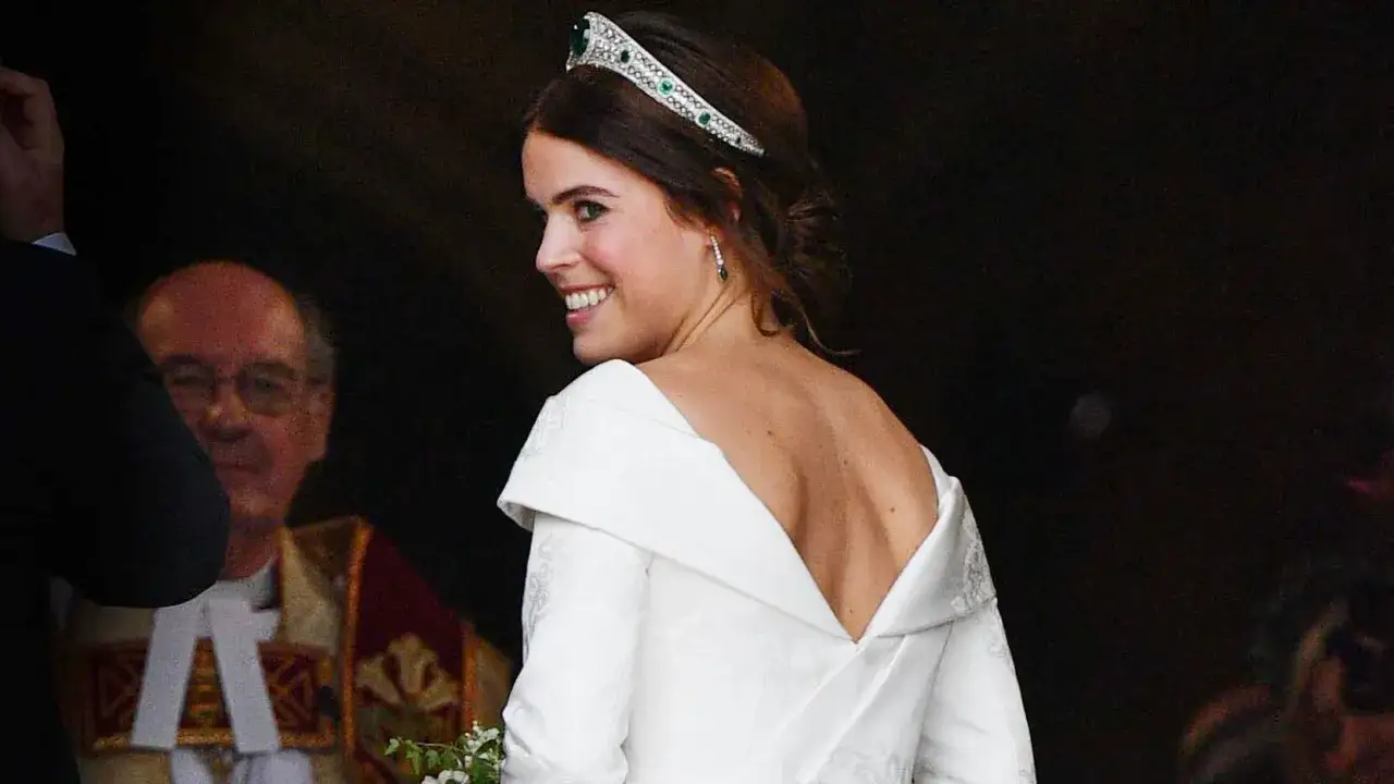 Prinsessan Eugenie ler glatt i sin br&ouml;llopskl&auml;nning med tiara. En man i pr&auml;stkl&auml;der syns i bakgrunden.