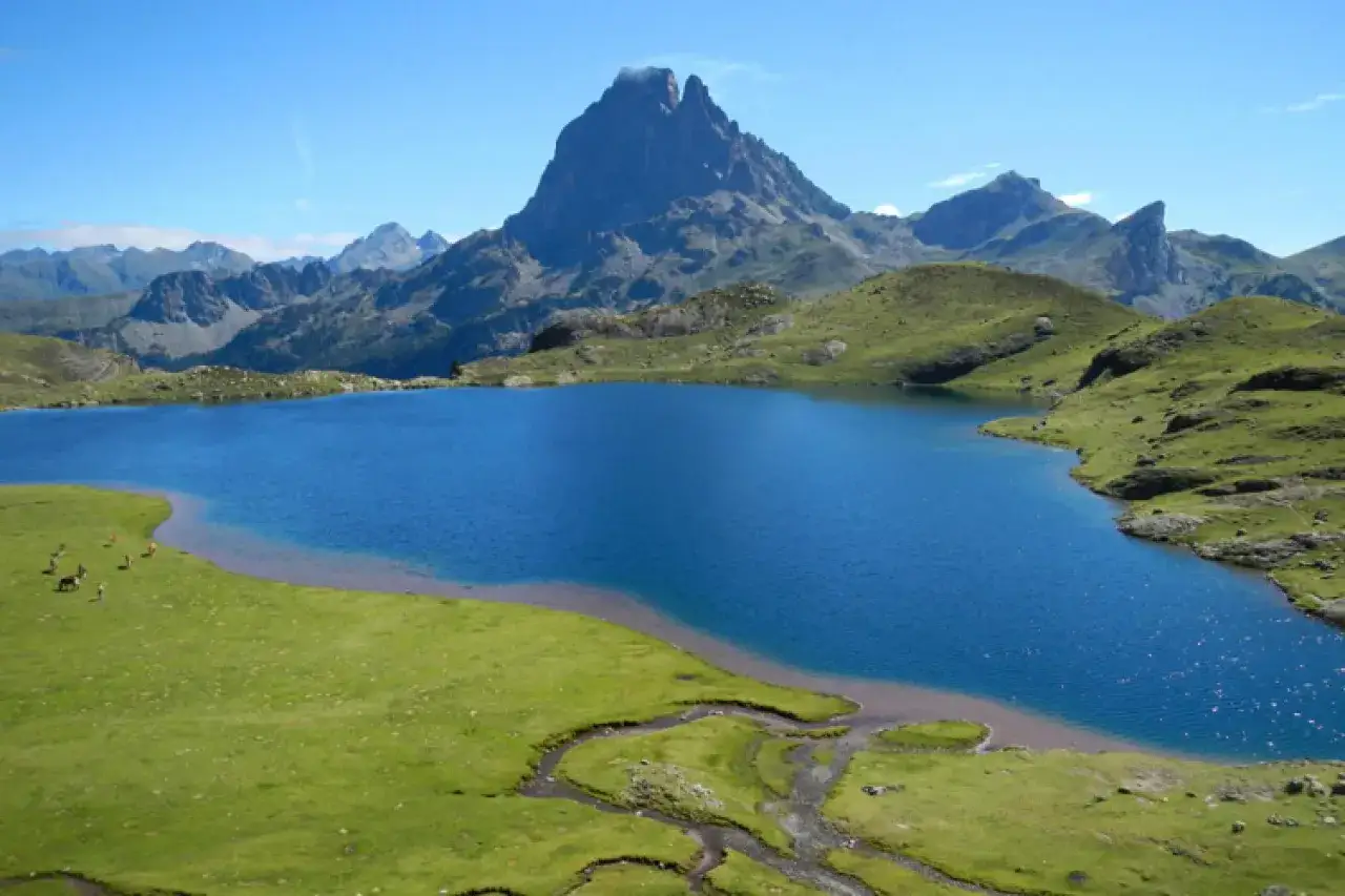 Lac bleu étincelant au **parque nacional de los Pirineos**, entouré de montagnes verdoyantes et d'un pic imposant. Quelques chevaux paissent paisiblement sur la rive.