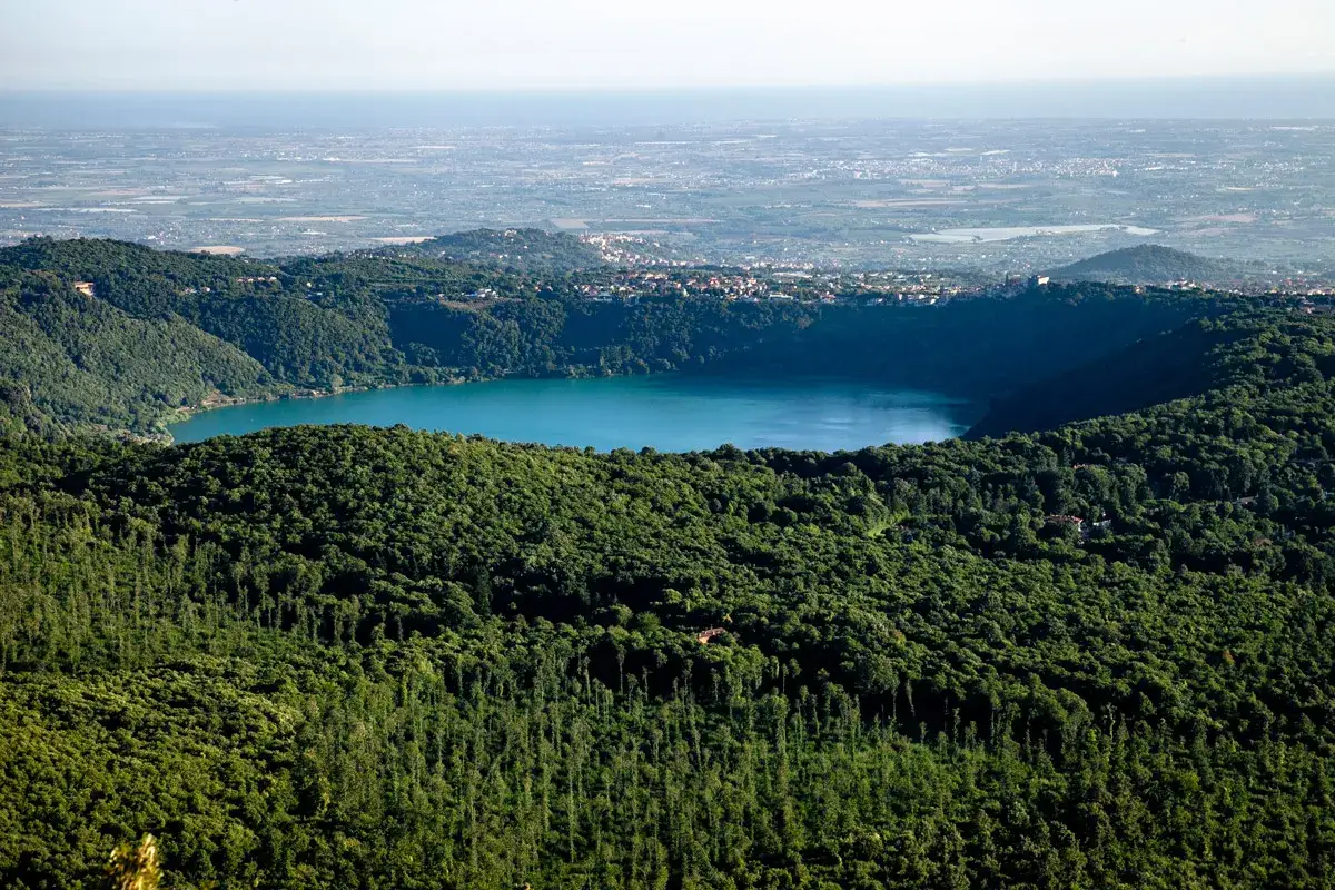 Vista panoramica del lago di Albano, incastonato tra i boschi lussureggianti, con il borgo di Rocca di Papa in lontananza.