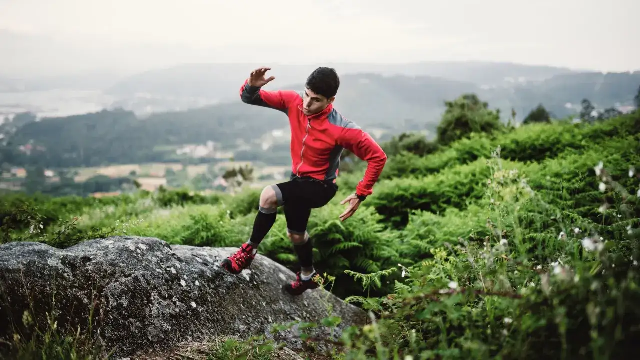 Un homme en tenue de sport rouge et noire fait du trail run sur un rocher, entouré de végétation luxuriante et d'une vue sur la vallée.