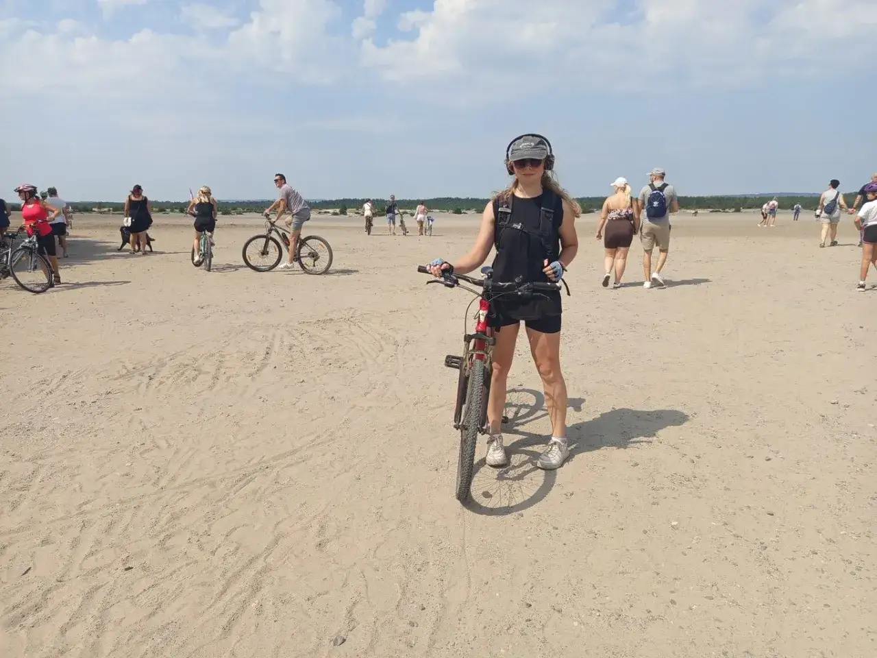 Dziewczyna z rowerem na piaszczystym terenie, gdzie inni też jeżdżą na rowerach i spacerują. To idealne miejsce na **velo przemsza**.