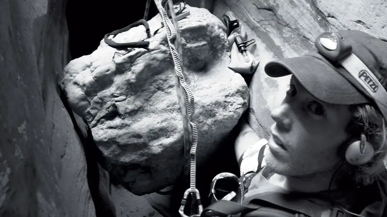 Aron Ralston, le visage marqué, est coincé dans un canyon, une corde à la main, face à un rocher menaçant.