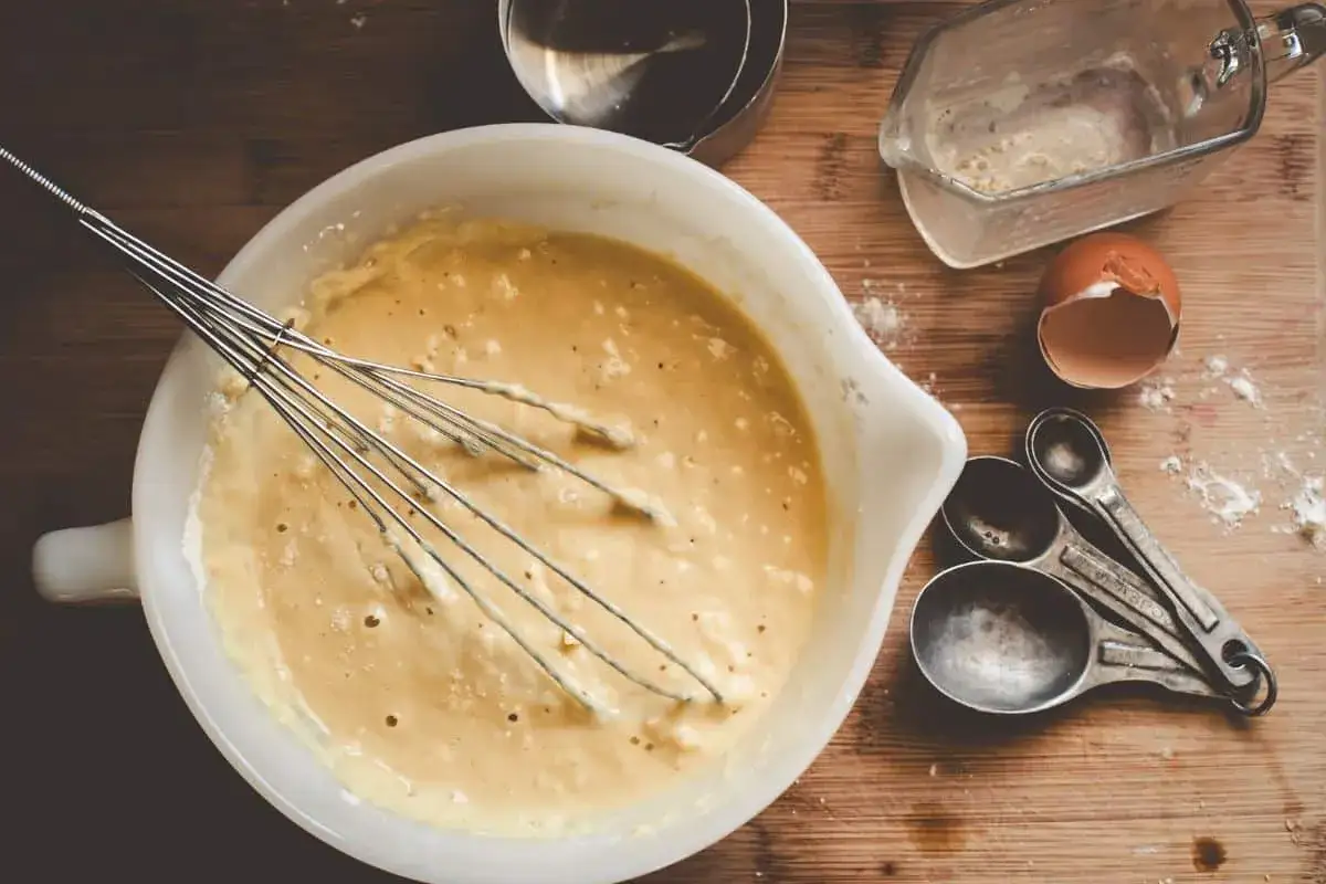 Pr&eacute;paration de cr&ecirc;pes &agrave; la farine de ch&acirc;taigne : p&acirc;te dans un bol avec un fouet, coquille d'&oelig;uf, cuill&egrave;res &agrave; mesurer et un pichet gradu&eacute;.