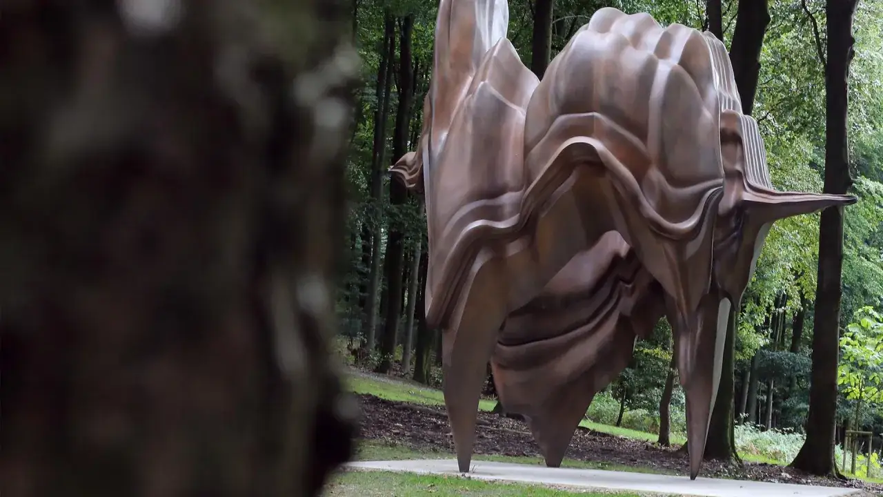 Eine organisch geformte, bronzefarbene Skulptur steht im **Skulpturenpark Waldfrieden** inmitten eines Waldes.