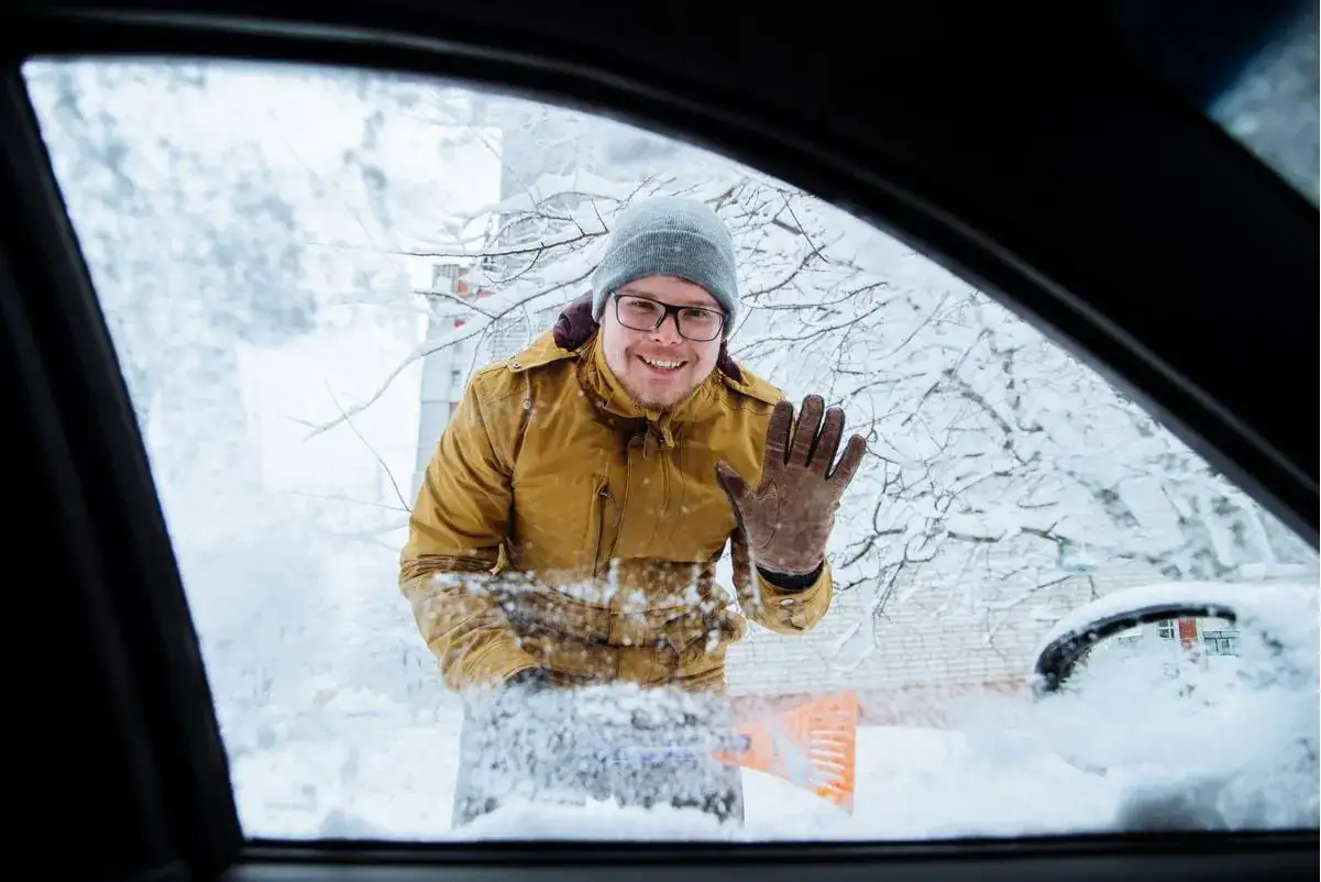 Glad man skrapar sn&ouml; fr&aring;n bilrutan f&ouml;r kallstart bil. Vinkar glatt trots kylan.