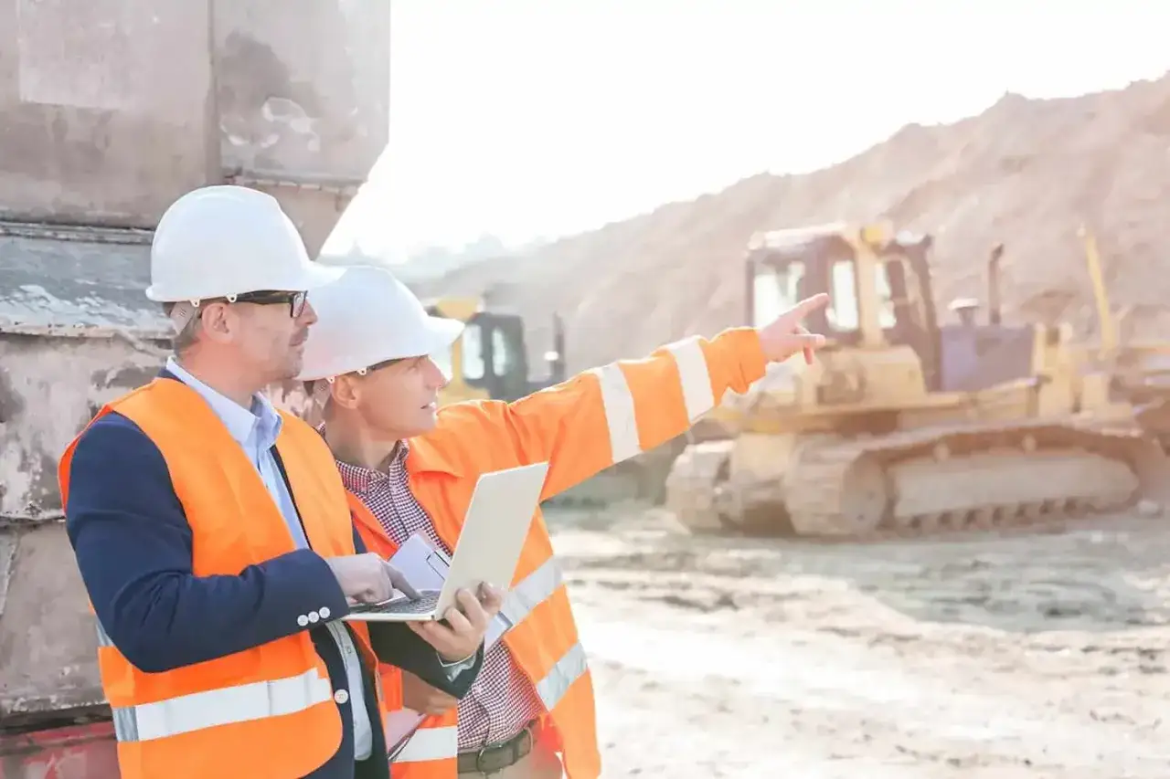 El director de obra y el director de ejecuci&oacute;n revisan planos en un port&aacute;til en una obra con maquinaria pesada.