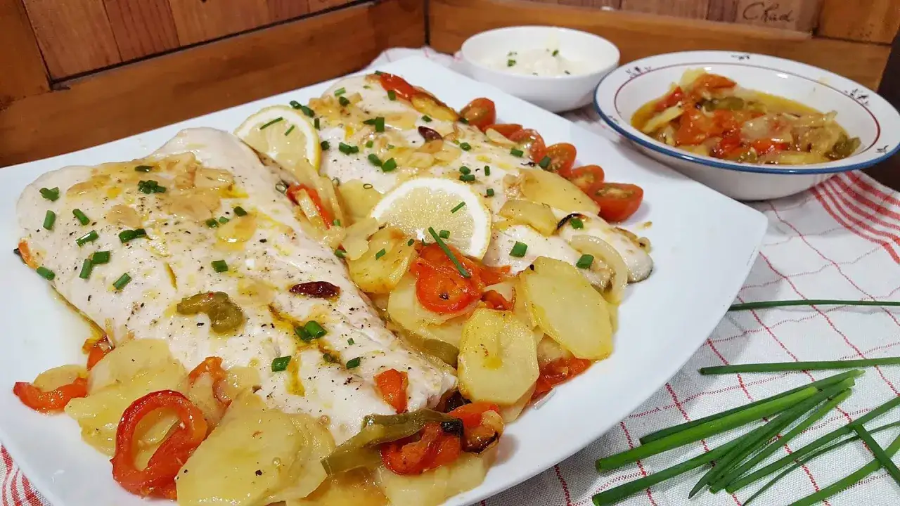 Deliciosa merluza al horno con patatas, pimientos, tomates cherry y rodajas de limón, todo espolvoreado con cebollino fresco.