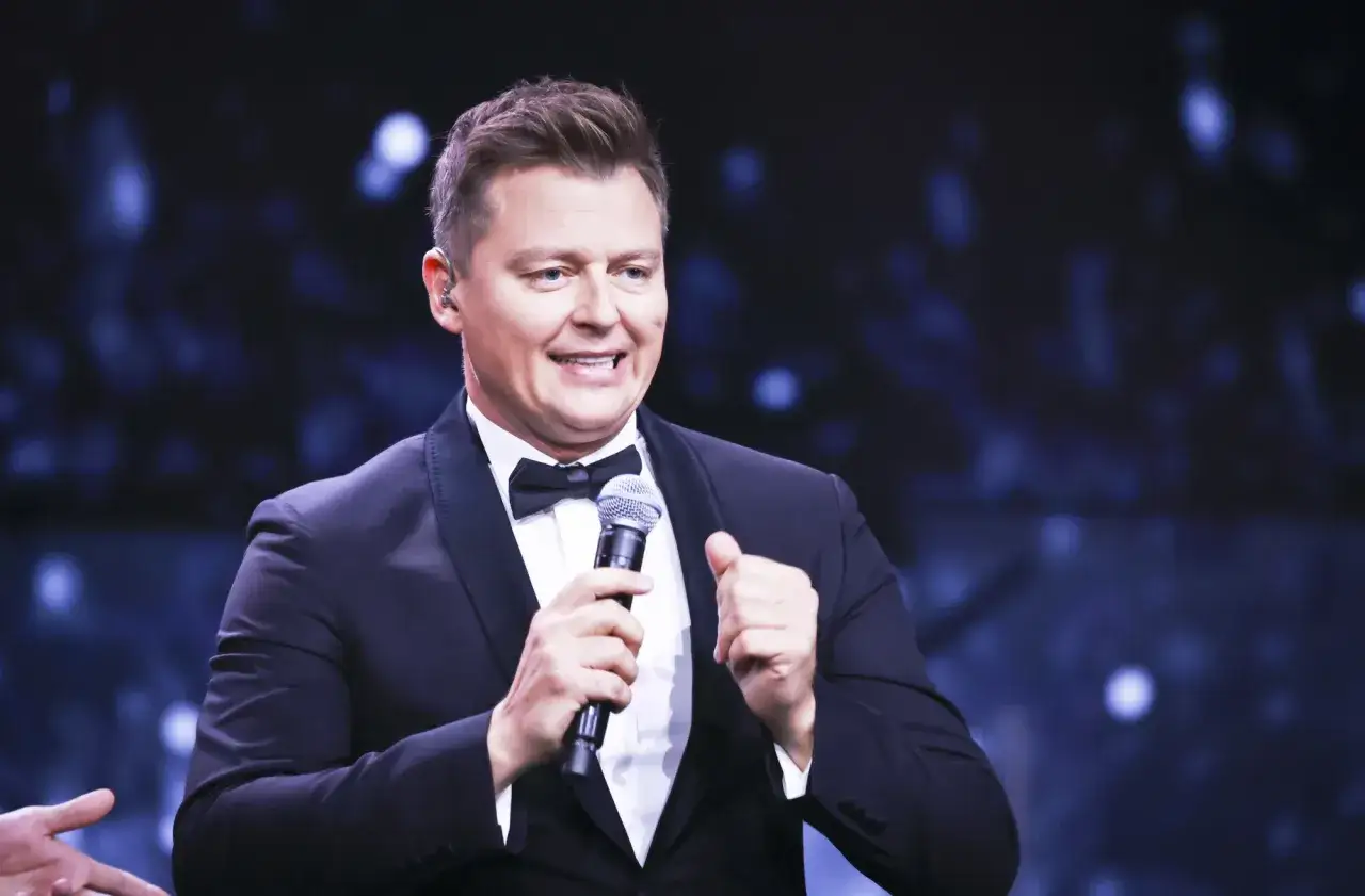 Rafał Brzozowski w garniturze z mikrofonem, uśmiechnięty. Czy to odpowiedź na pytanie dlaczego Rafał Brzozowski odszedł z Koła Fortuny?